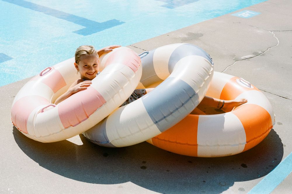Colorful pool floats in teal, pink, white and orange arranged on a concrete surface near water.