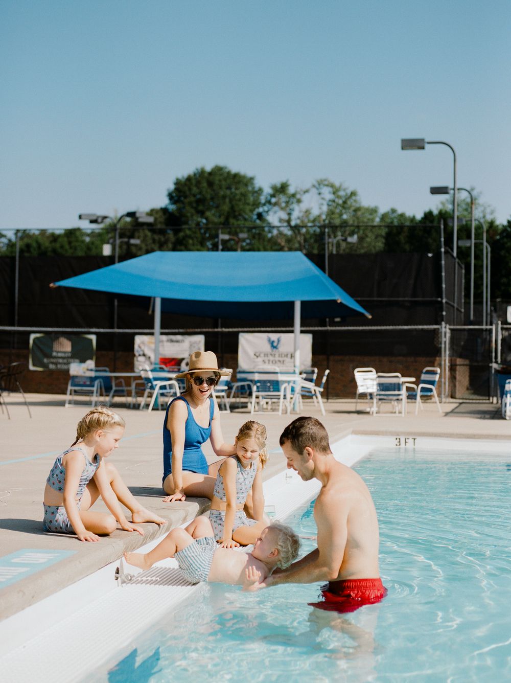 People gathering at a community pool under a blue shade canopy on a sunny day.