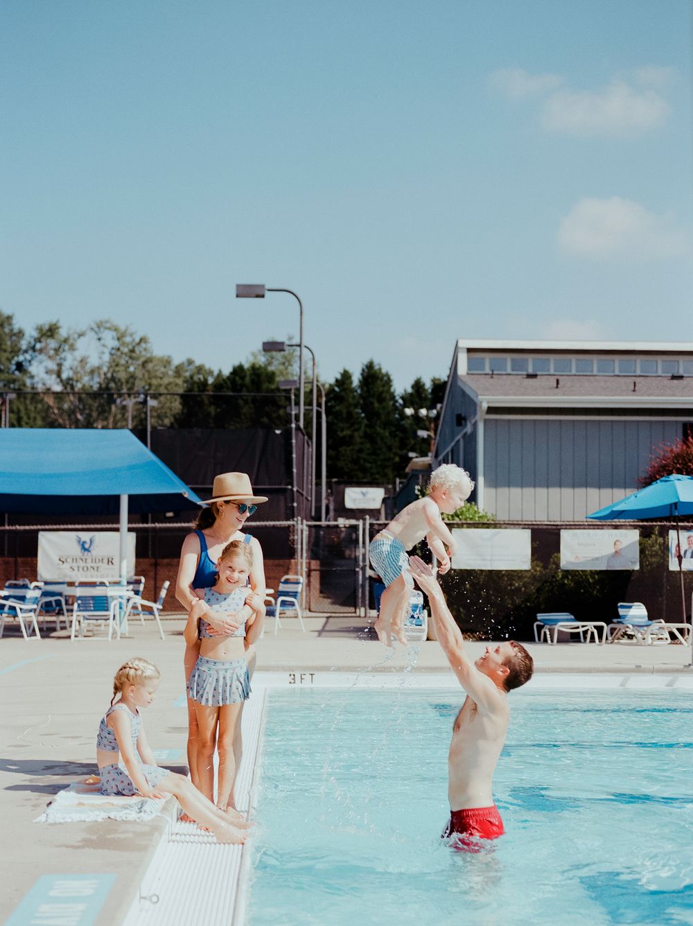Summer pool scene with people playing and swimming under blue canopy shade structures.