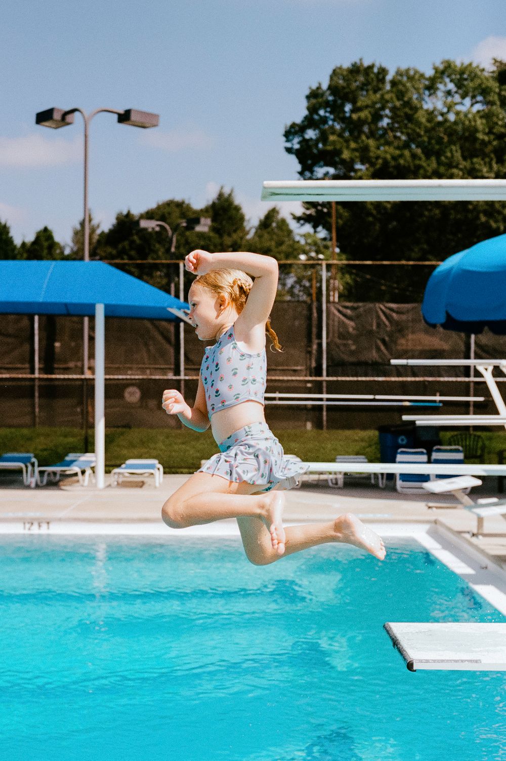Someone wearing floral swimwear joyfully leaps into a bright blue swimming pool on a sunny summer day.