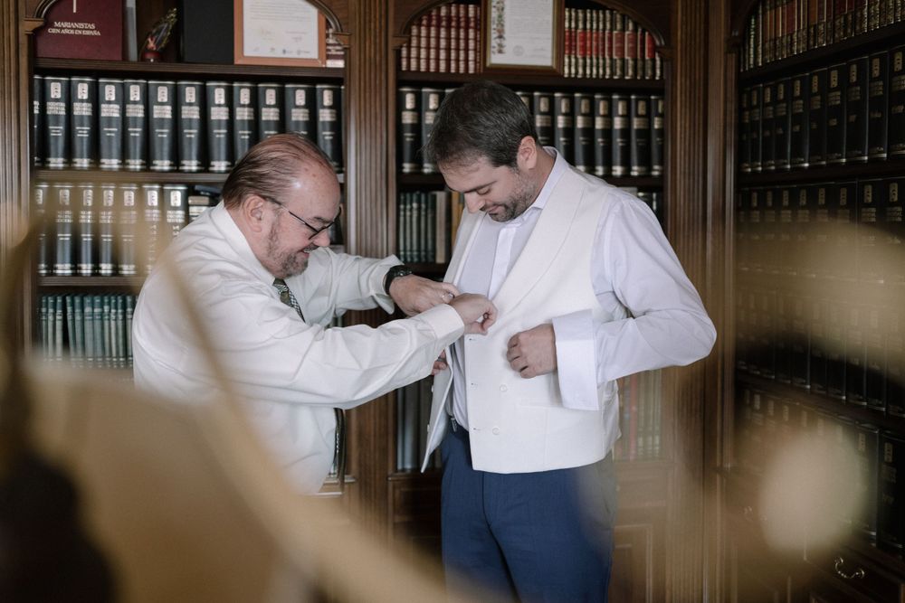Two people in white shirts adjusting formal attire in a library or office setting with bookshelves in background.