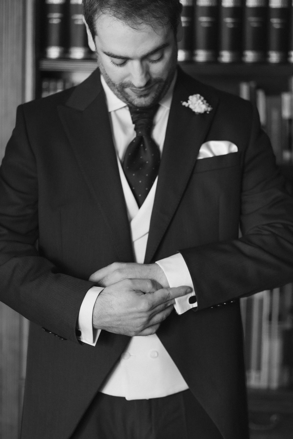 Black and white photo of groom in formal tuxedo adjusting his cufflink while smiling, with bookshelves in background.