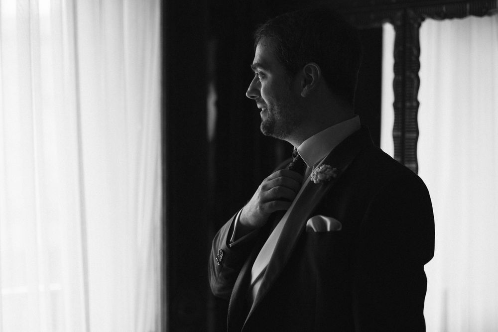 A black and white portrait of a man in a suit adjusting his tie near a window with dramatic side lighting.