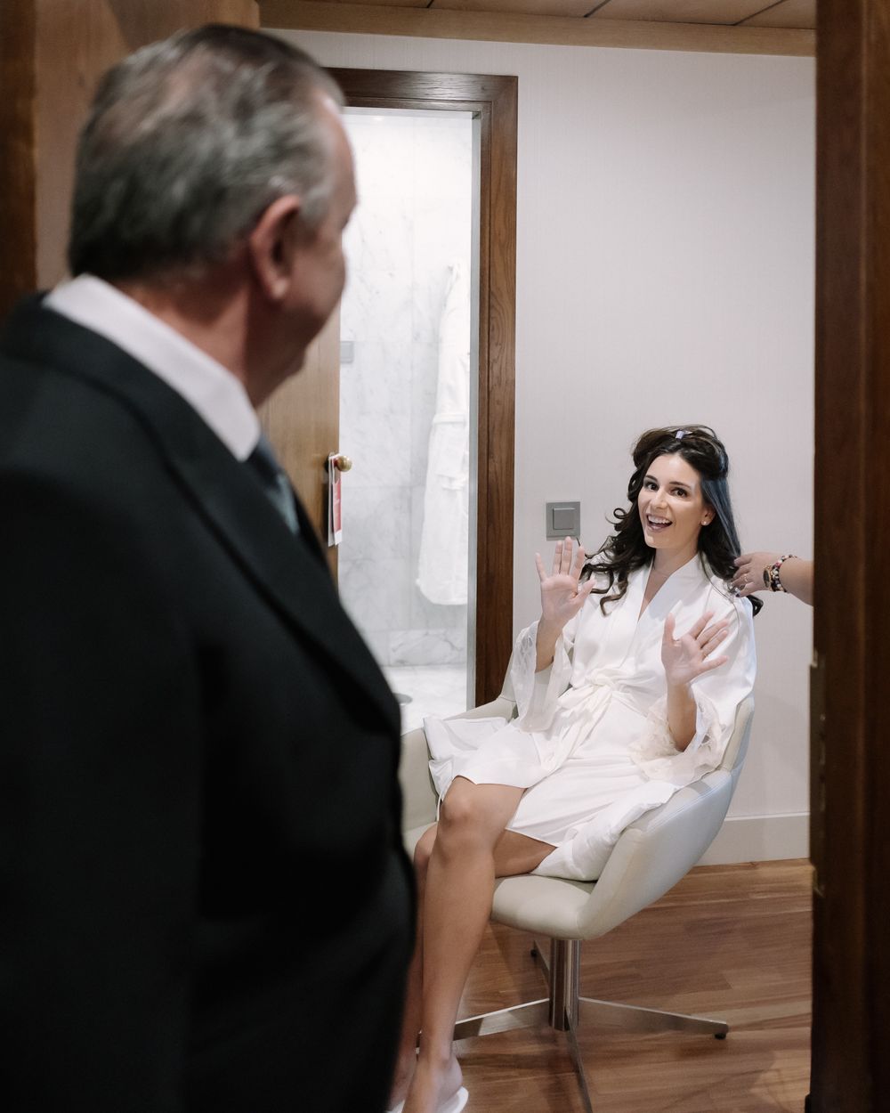 A woman wearing a white silk robe sits in a modern chair while a man in a suit stands in the doorway of a room.