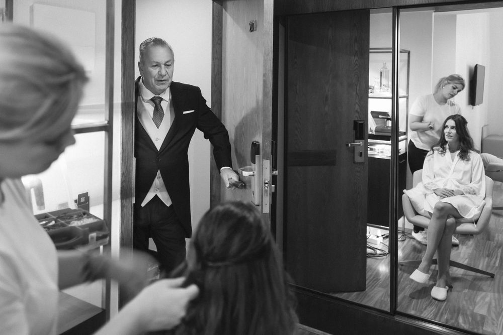 Black and white photo showing a groom in formal attire waiting while others prepare in the dressing room.