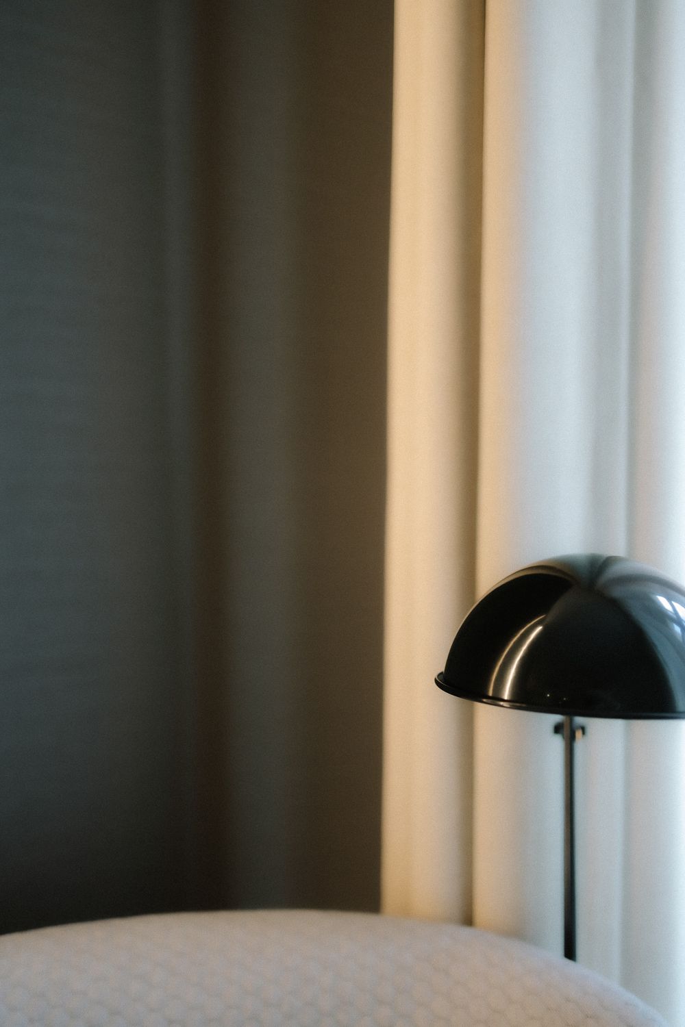 A black dome desk lamp sits beside white curtains and wall, creating a minimalist interior aesthetic.