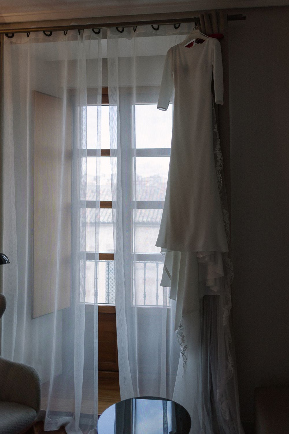 A white wedding dress hangs elegantly by a sunlit window with sheer curtains in a bridal preparation room.