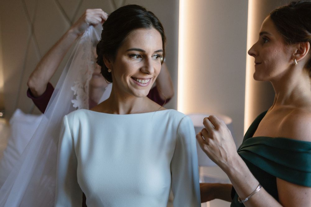 A bride in a sleek white bateau neck wedding dress smiles while getting ready with help adjusting the veil.