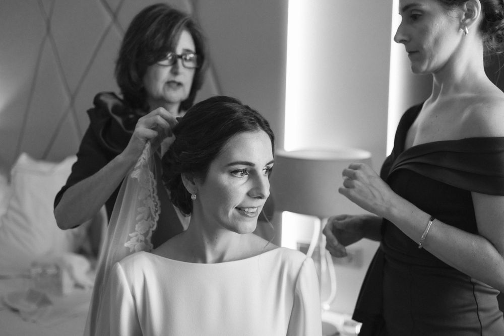 A black and white photo shows bridal preparations with the bride in a white dress getting her hair styled while others assist.
