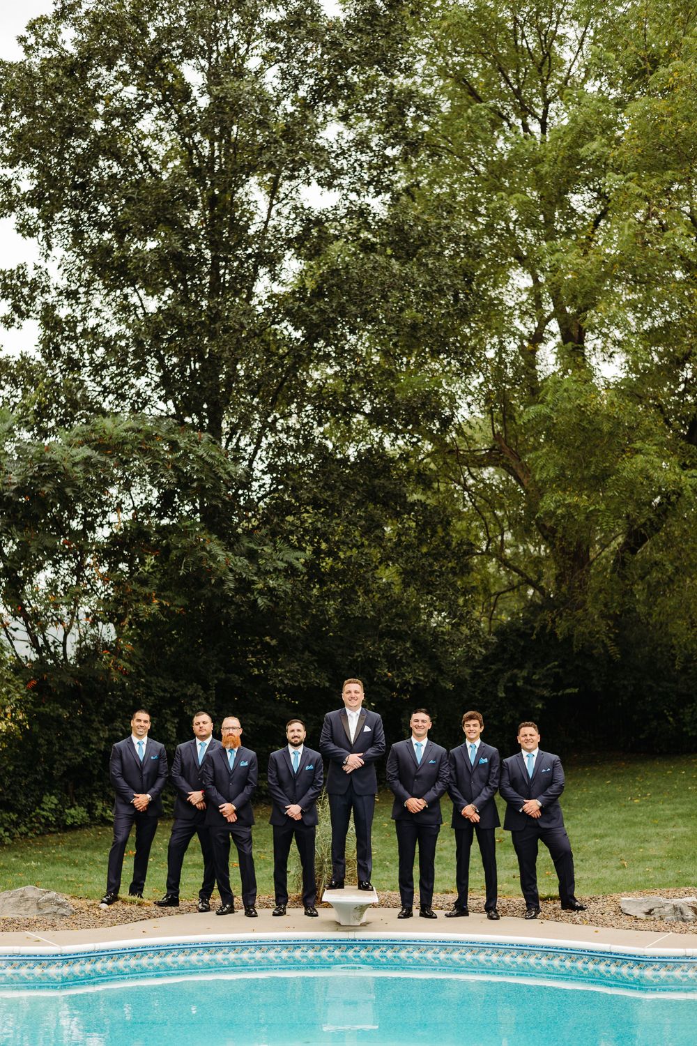Groomsmen lined up in navy suits along curved edge of swimming pool during outdoor wedding photo session.