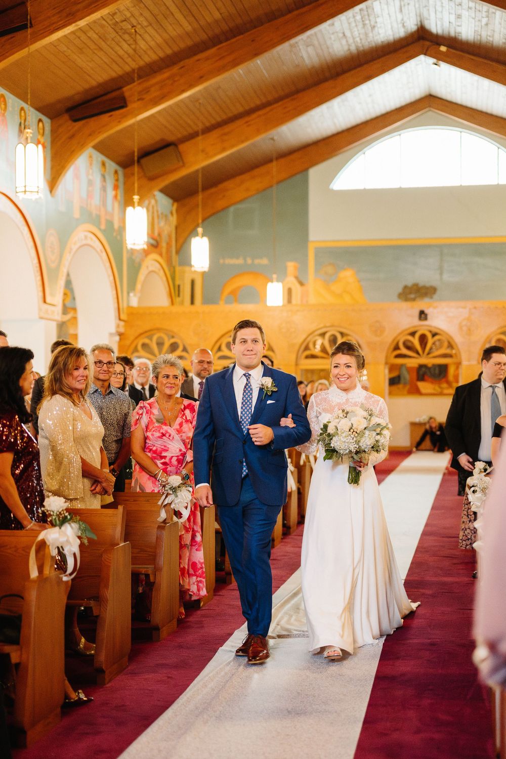 Wedding procession walks down a red-carpeted church aisle with wooden beams overhead and arched stained glass windows.