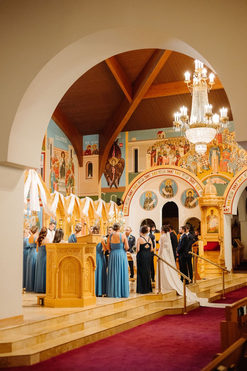 Wedding party lined up along ornate church altar steps with elegant crystal chandeliers and golden decor.