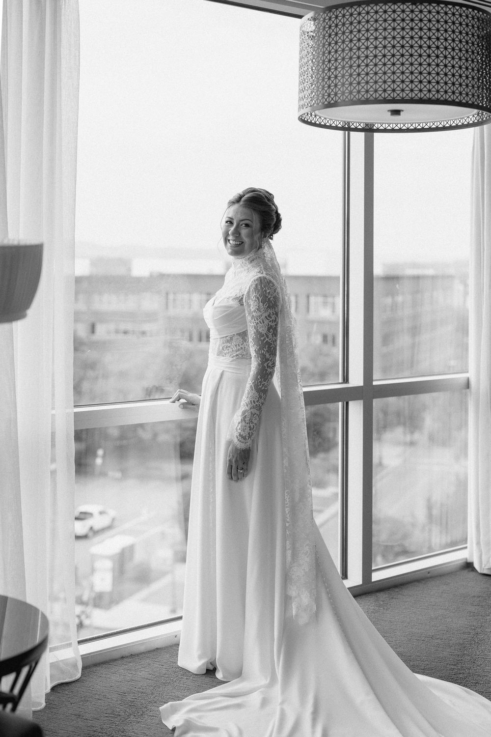 A person in a flowing white wedding dress stands by large windows in a bright modern interior space.