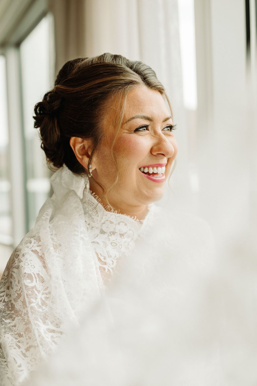 A bride in a white lace wedding dress smiles joyfully while posing near bright windows.