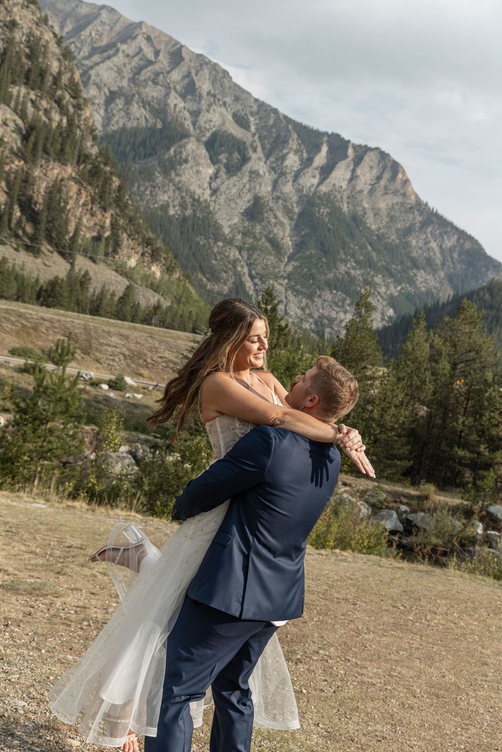 A couple embraces on a mountain ridge with dramatic rocky peaks towering in the background under cloudy skies.