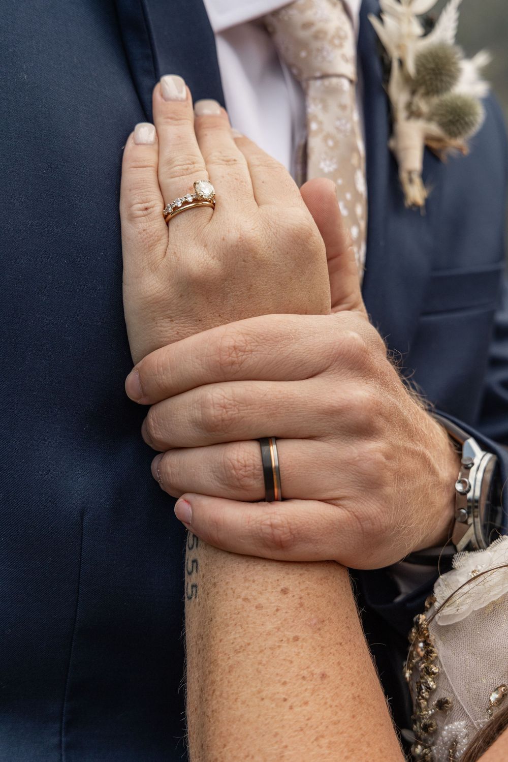 Close up view of hands wearing wedding rings, showing intimate embrace against dark clothing.