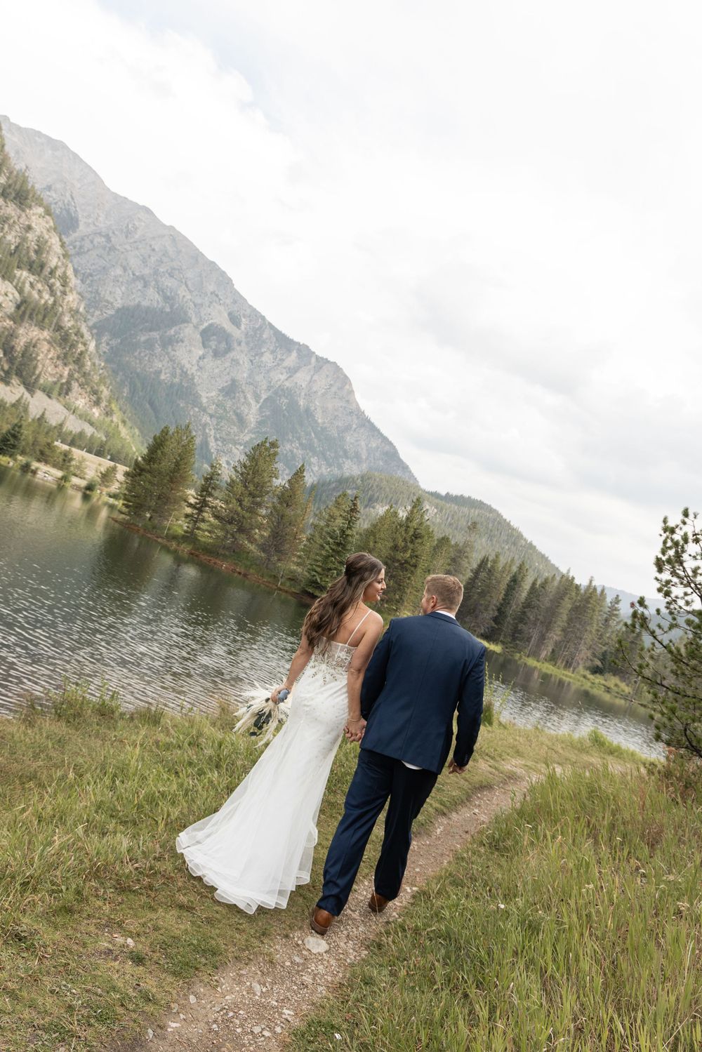 Couple walking along scenic mountain lake path in romantic wedding attire with dramatic peaks in background.