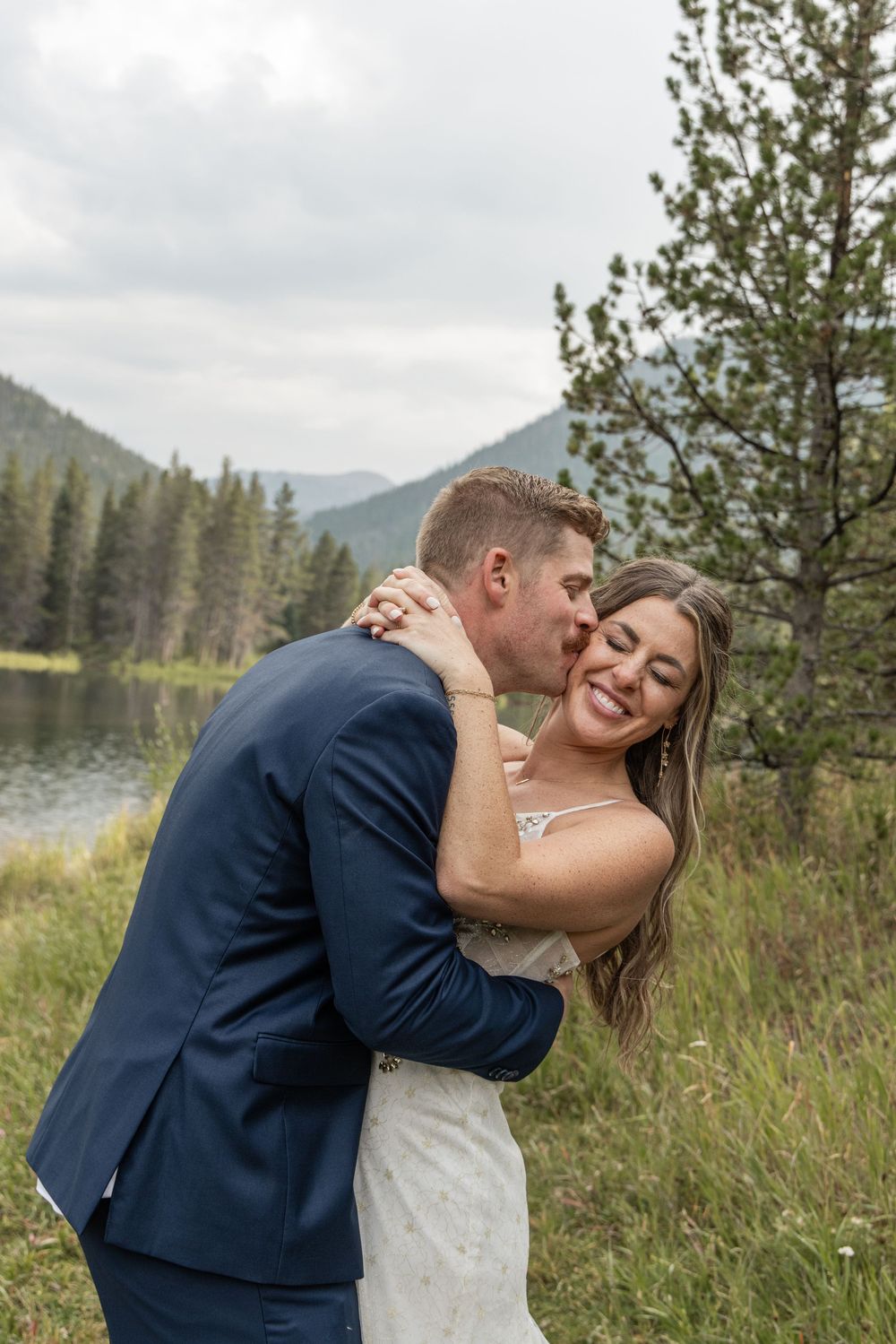 A romantic couple in wedding attire embrace and share a kiss in front of a scenic mountain landscape with pine trees.