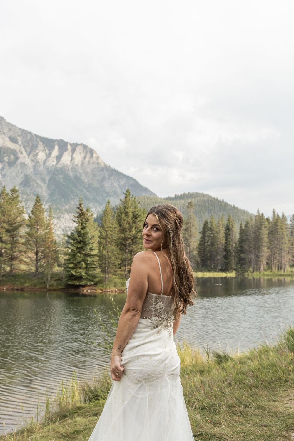 A bride in a flowing white dress stands at the edge of a mountain lake surrounded by evergreen trees and dramatic peaks.