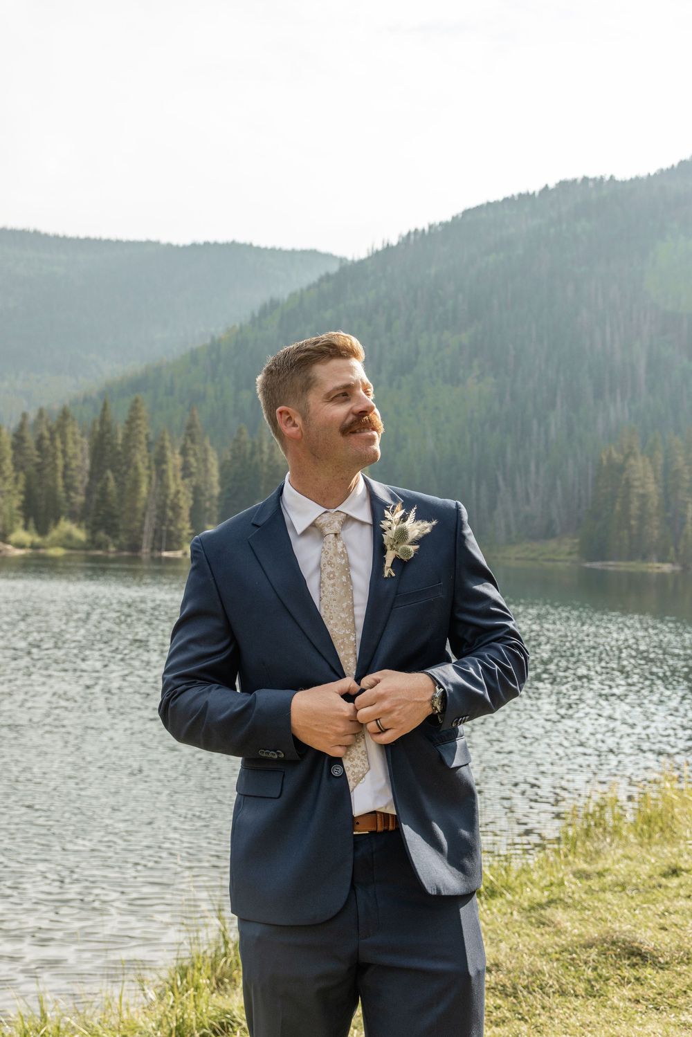 A groom in a navy suit stands by a scenic mountain lake surrounded by evergreen trees and hills.