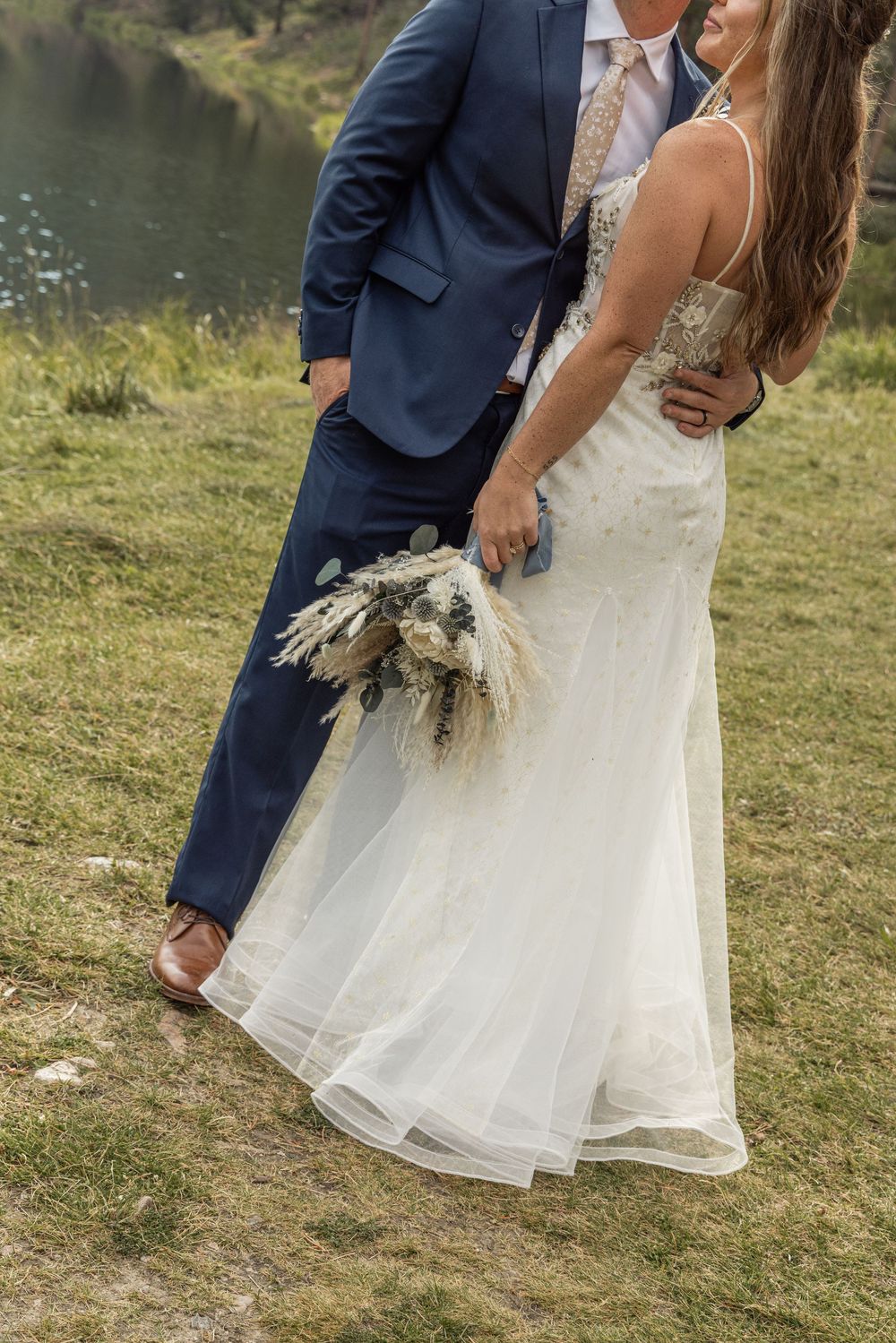Close-up collage of a bride's lace wedding dress and groom's navy suit captured near a lake setting at sunset.