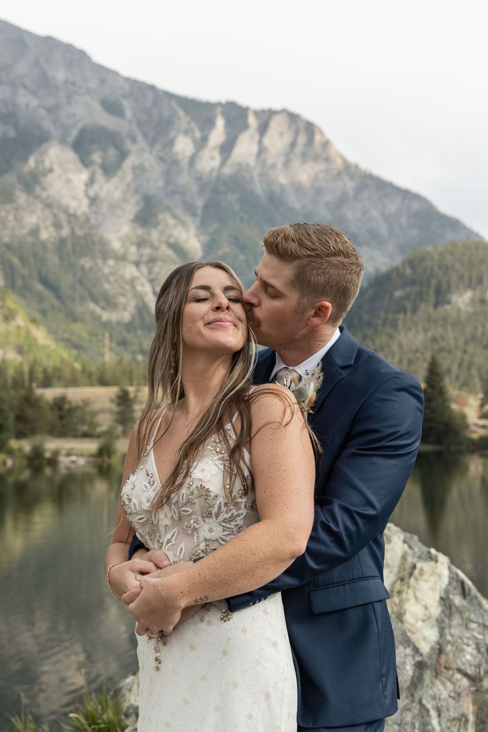 Romantic couple embraces by a scenic mountain lake with rocky cliffs in the background.