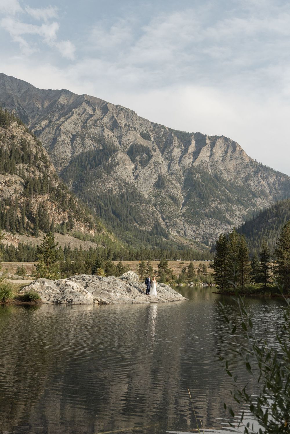Mountain range reflects in tranquil lake with rocky shoreline and evergreen trees under cloudy skies at Banff National Park.