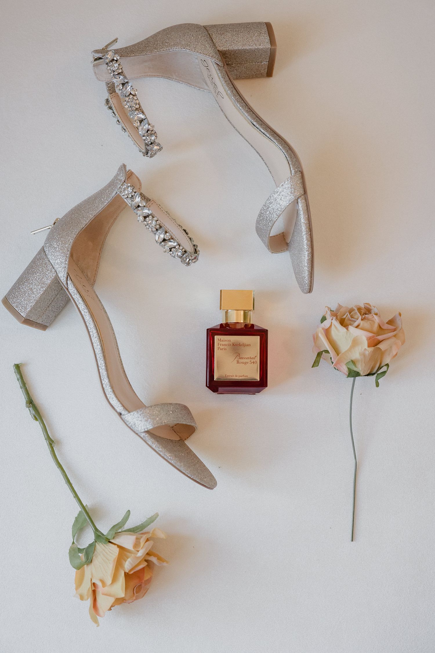 Wedding accessories flatlay with sparkly heels, perfume bottle and peach roses on white background.