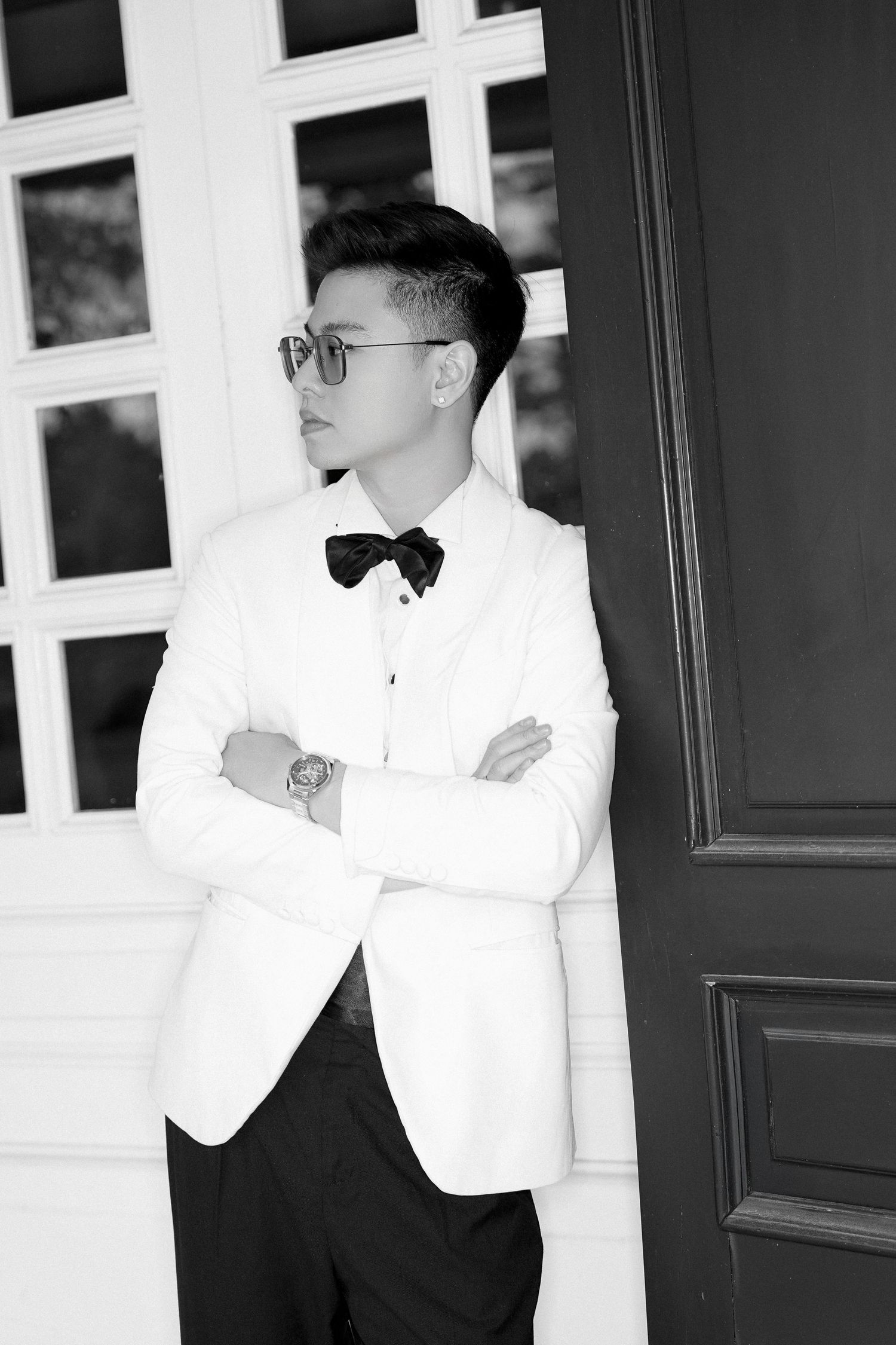 Elegant Asian groom in white tuxedo with bow tie and sunglasses posing with crossed arms in black and white.