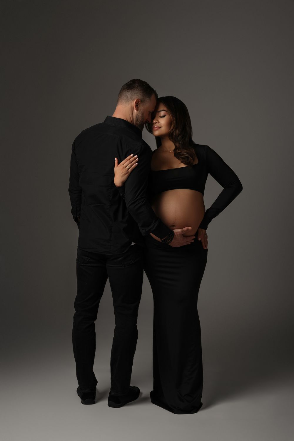 Couple dressed in all black embrace tenderly in a professional studio maternity photography session.