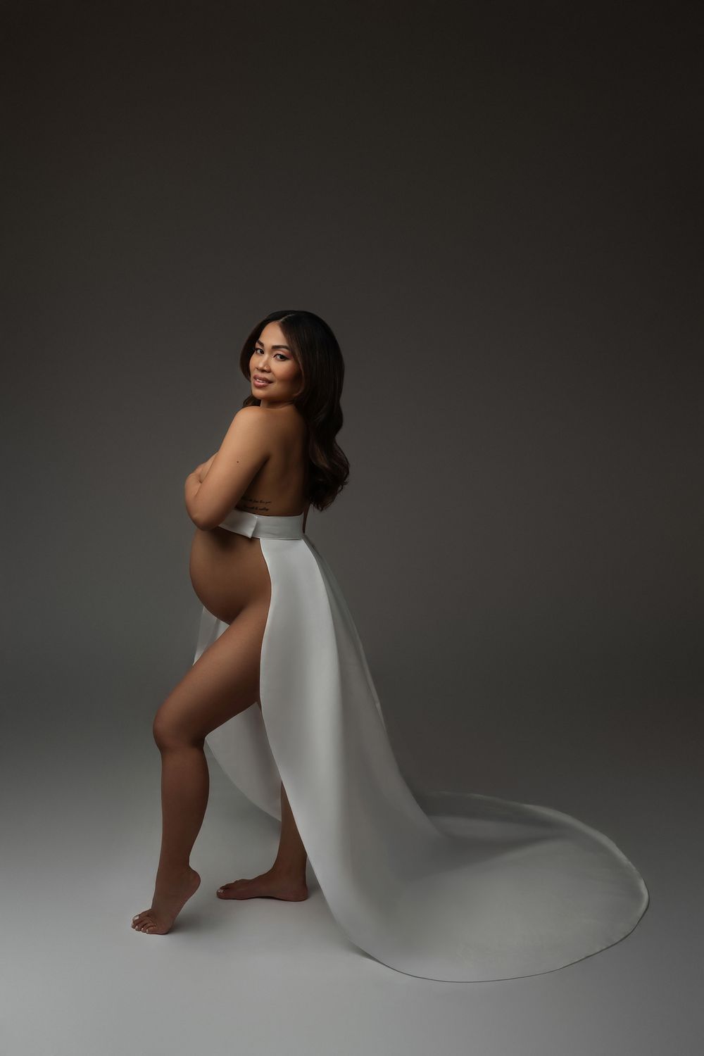 Pregnant woman in a white draped fabric poses sideways, showcasing her baby bump in a studio maternity photo.