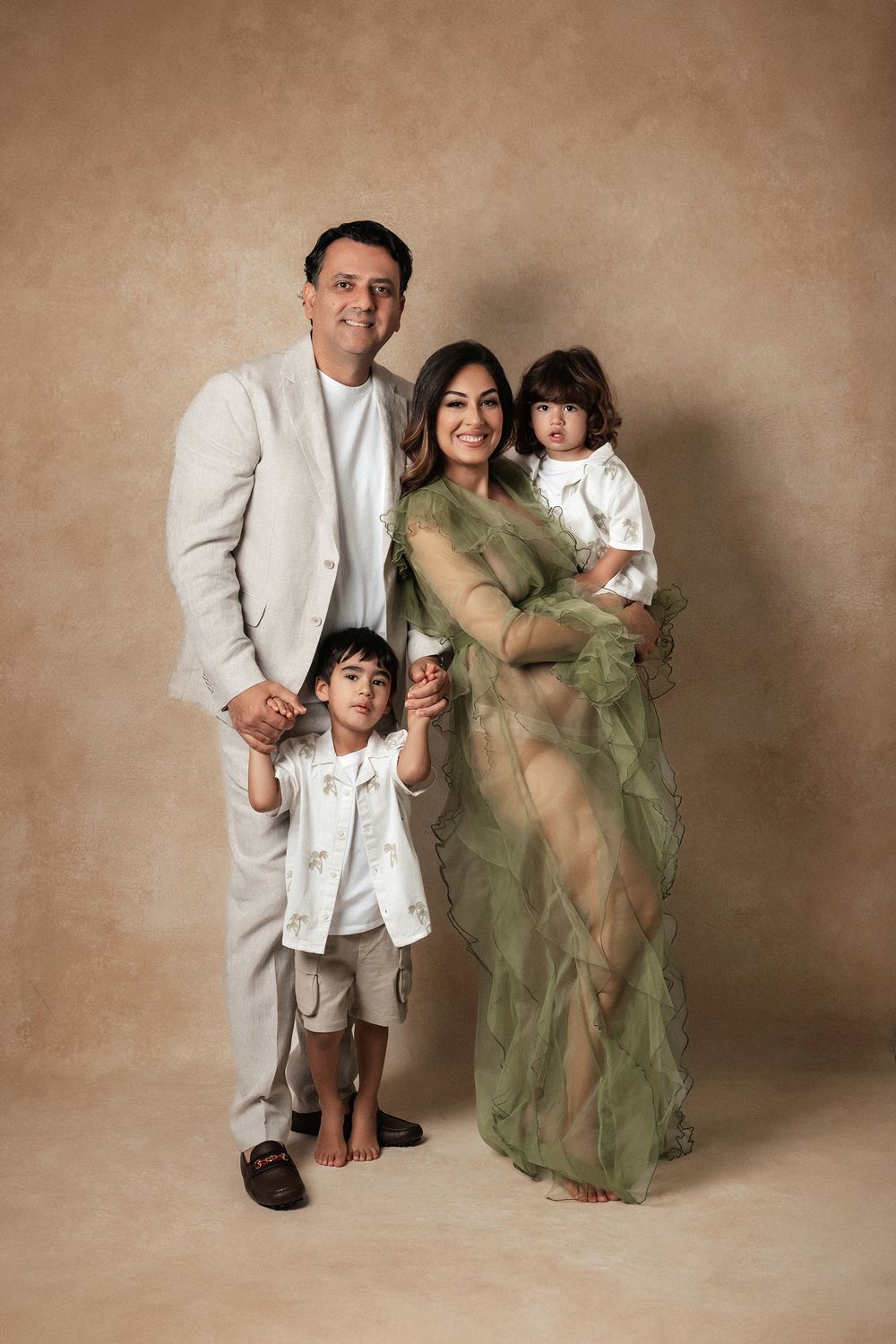 Family of four poses together with pregnant mother in a green tulle gown for a studio maternity portrait.