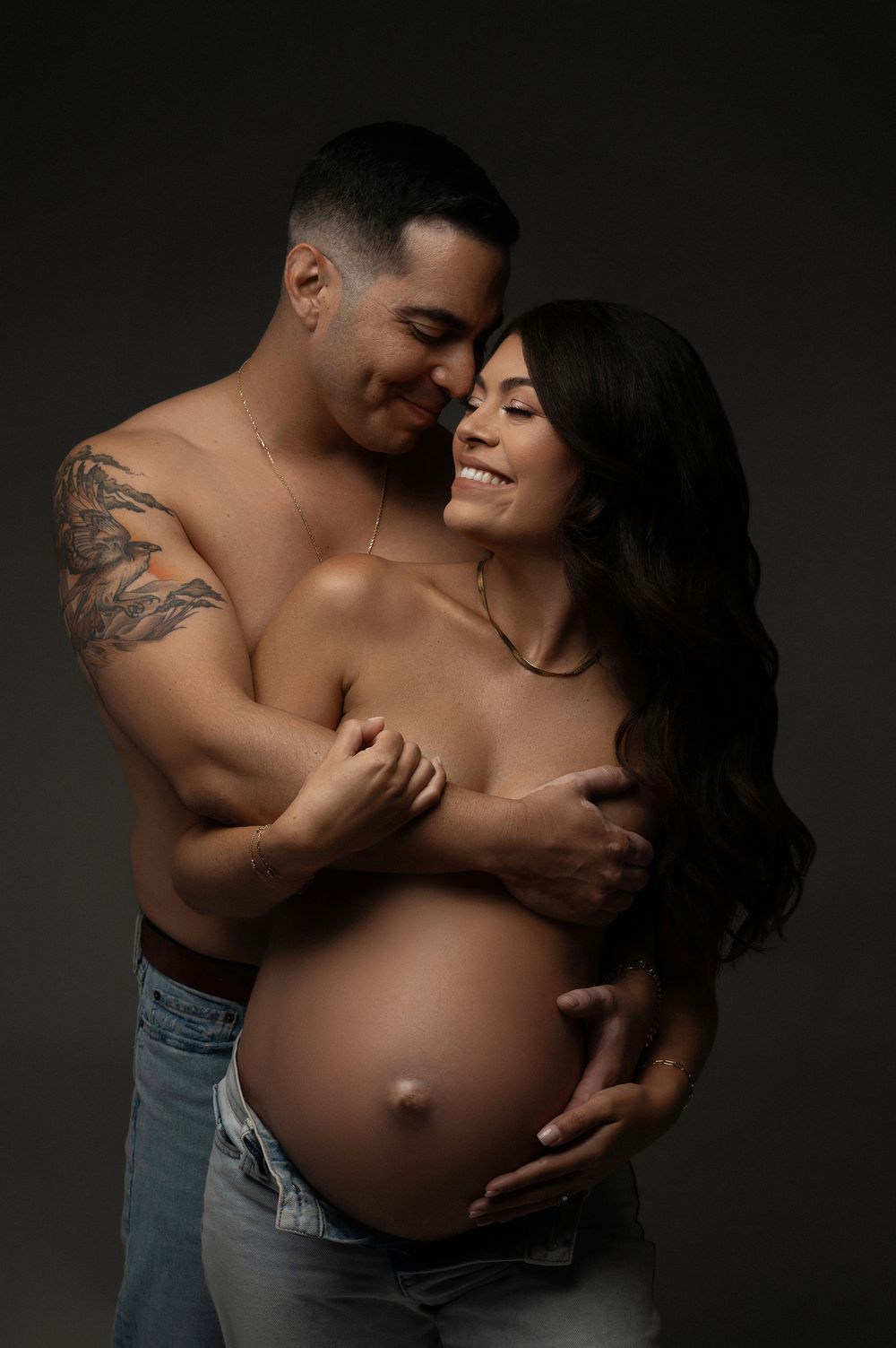 Shirtless tattooed man embraces smiling pregnant woman in a tender studio couples maternity photography session.
