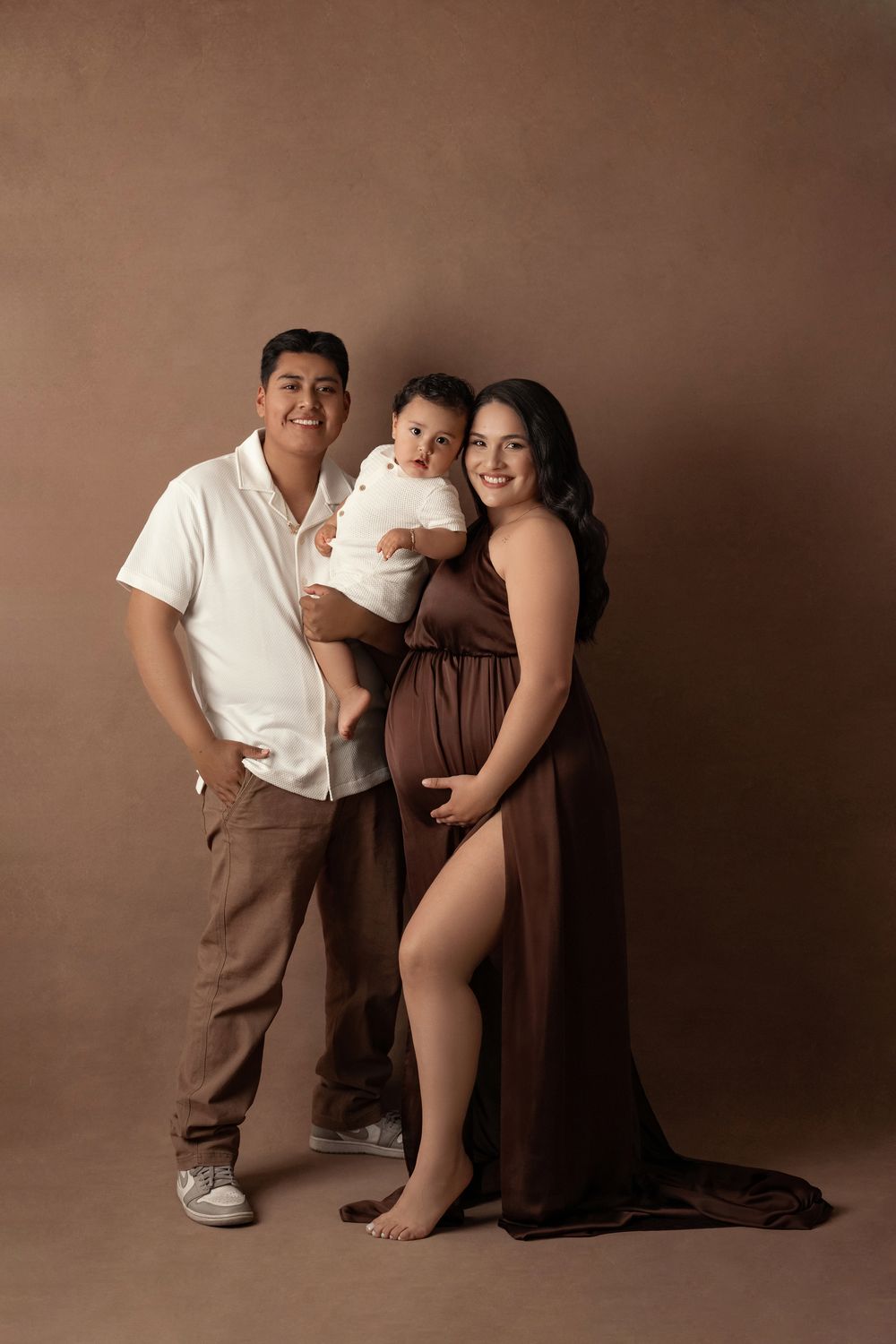 A smiling pregnant woman in a brown gown poses with her partner and baby in a studio.