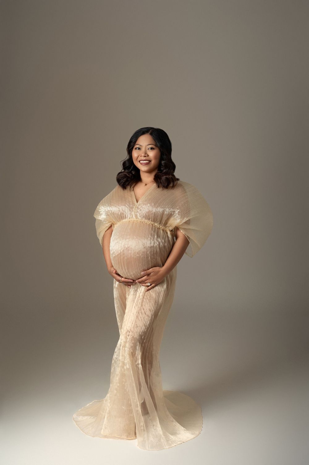 Smiling pregnant woman in gold sequin tulle maternity gown poses in professional photo studio.
