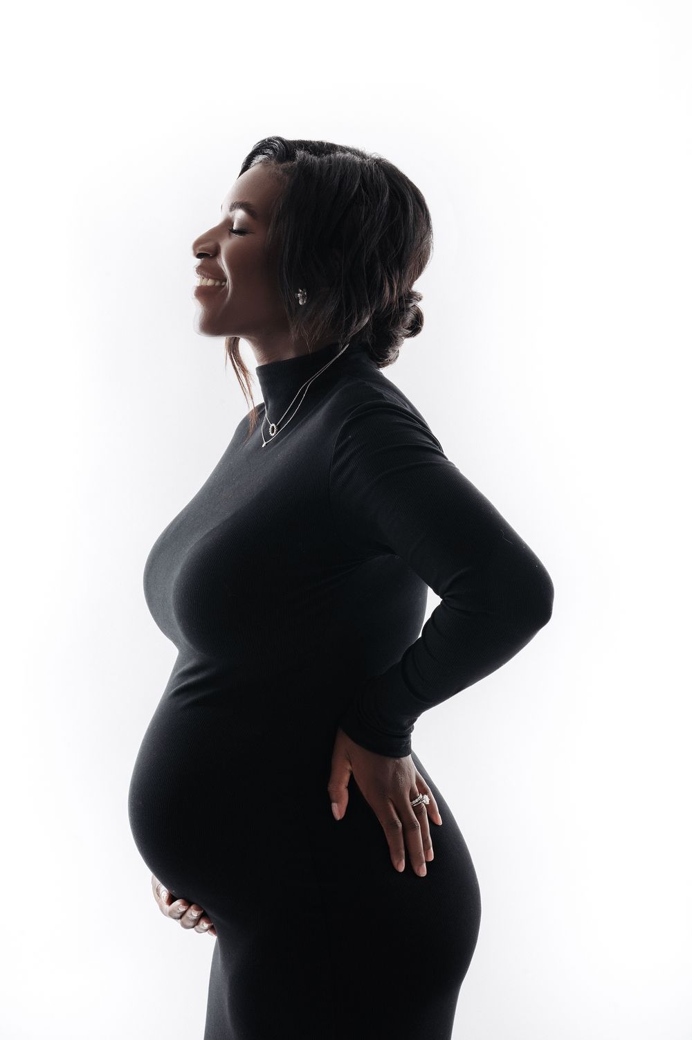 Pregnant woman in black turtleneck dress poses in striking backlit silhouette profile shot.