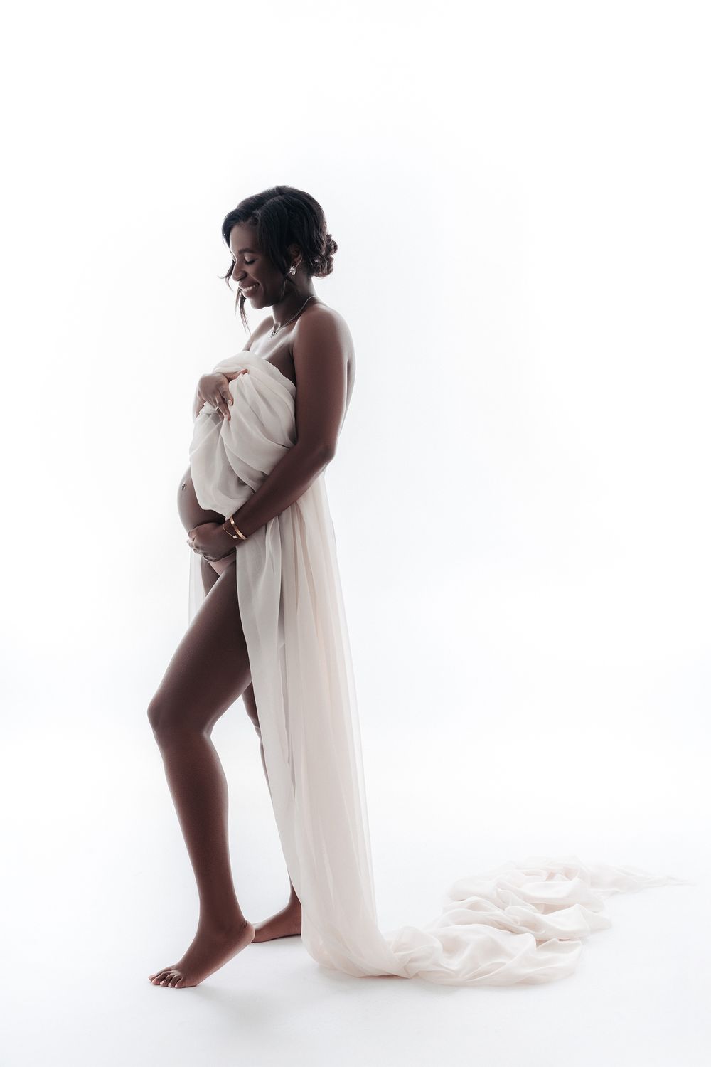 Pregnant woman wrapped in flowing white fabric poses in elegant backlit studio portrait.
