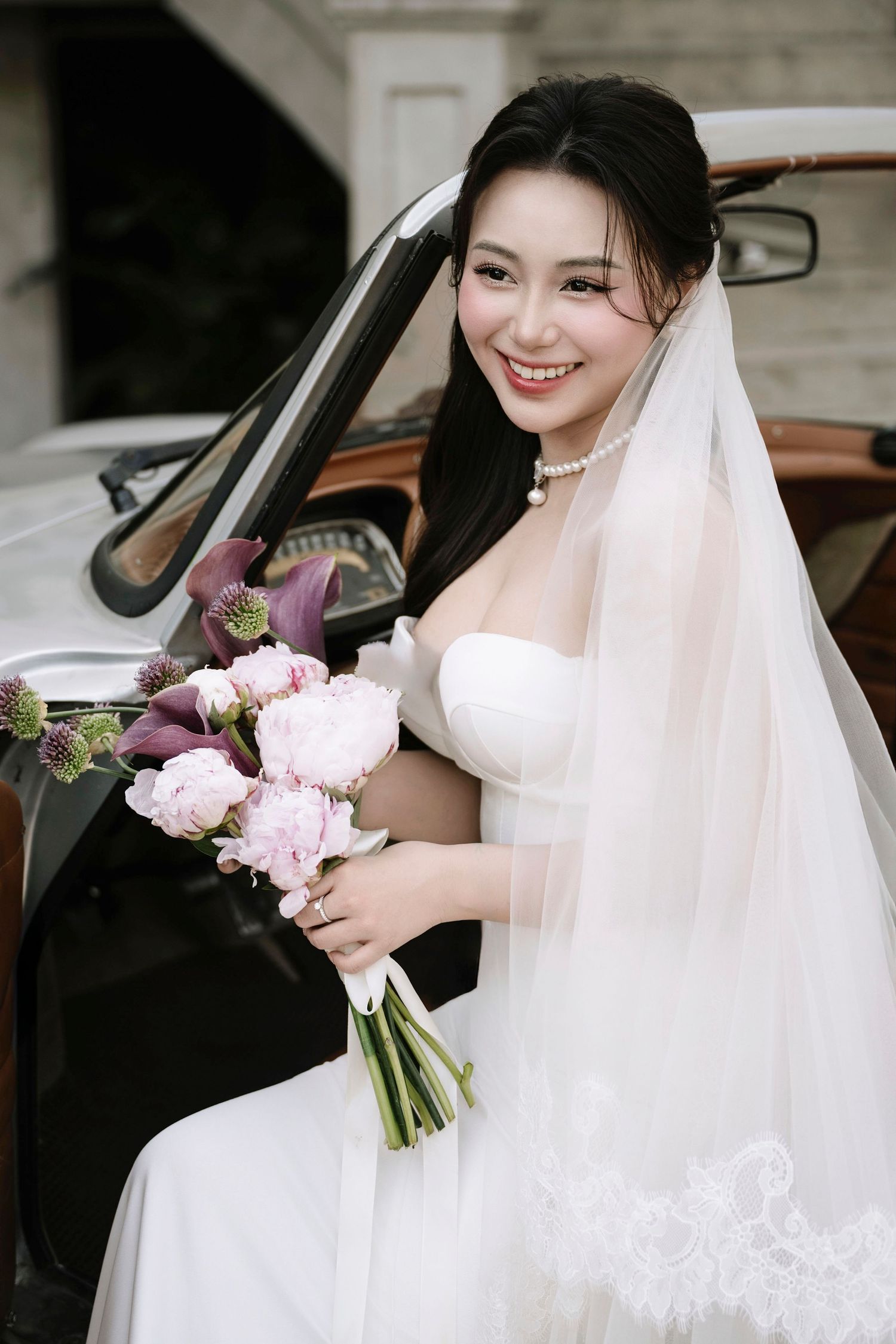 Smiling Asian bride in white strapless gown and veil holds pink bouquet beside vintage classic car.