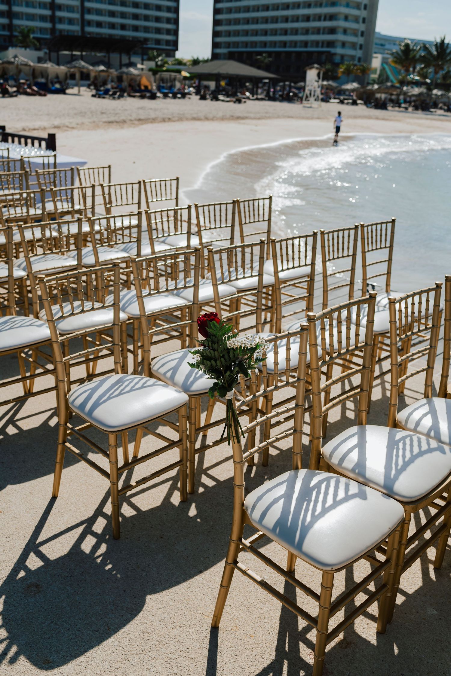 Rows of wooden chiavari chairs with white cushions arranged for a beach wedding ceremony under bright sunlight.