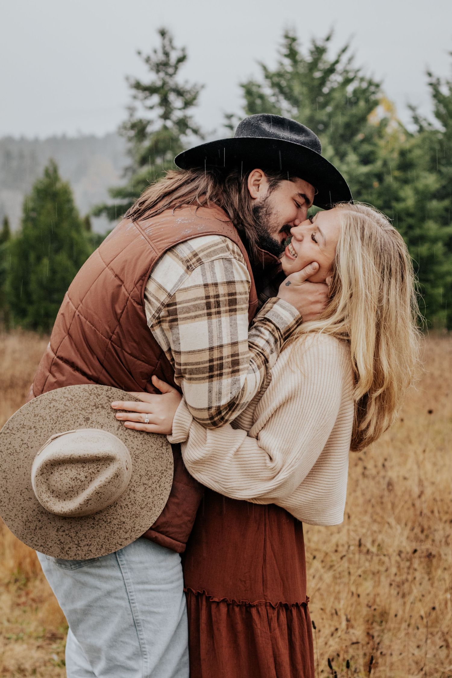 Couple embracing tenderly in a rainy field, both wearing cowboy hats and autumn-toned outfits.
