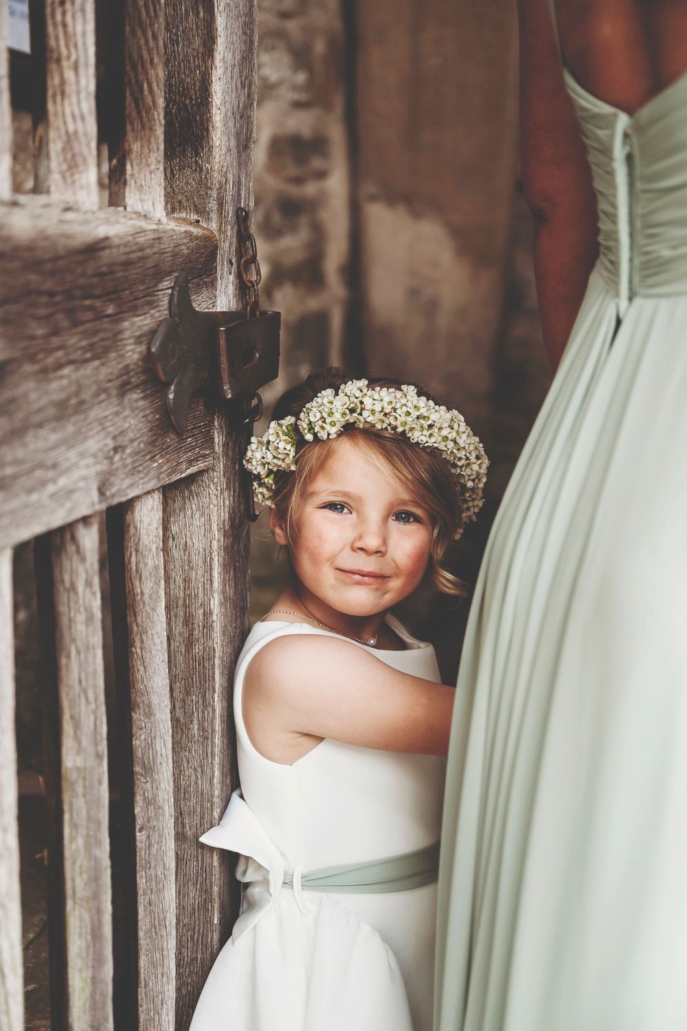 Small flower girl in white dress embracing dress fabric among rustic wooden beams.