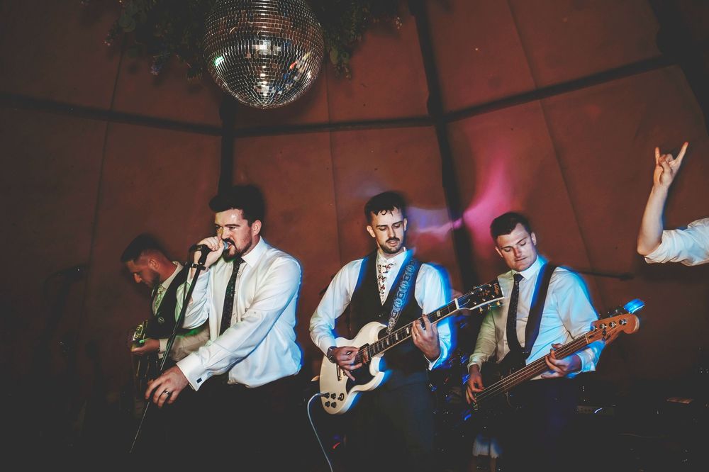 Band performing on stage with multiple musicians in white shirts and ties under a disco ball.