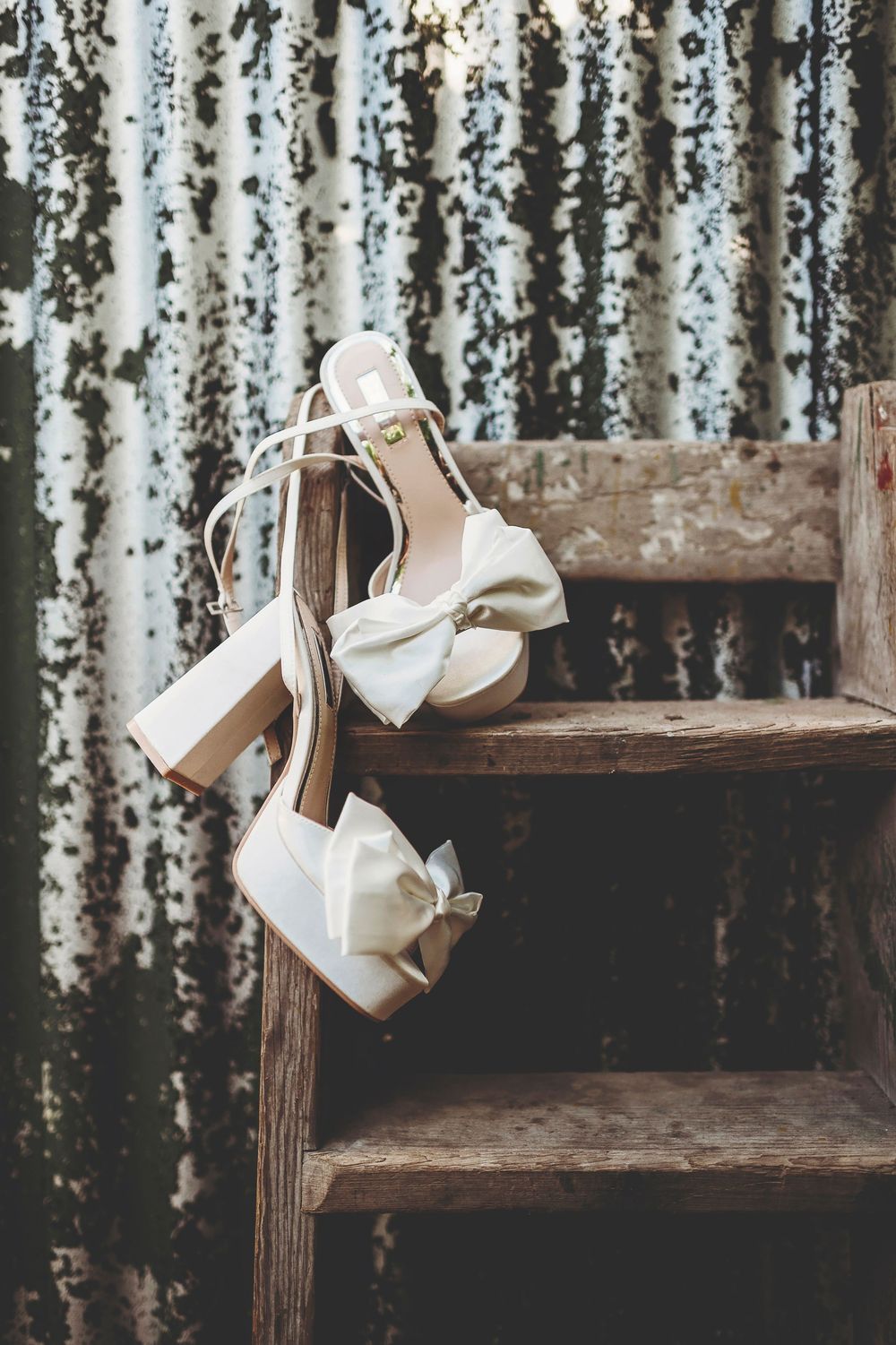 White bridal heels with bows hanging on rustic wooden stairs against a weathered metal background.
