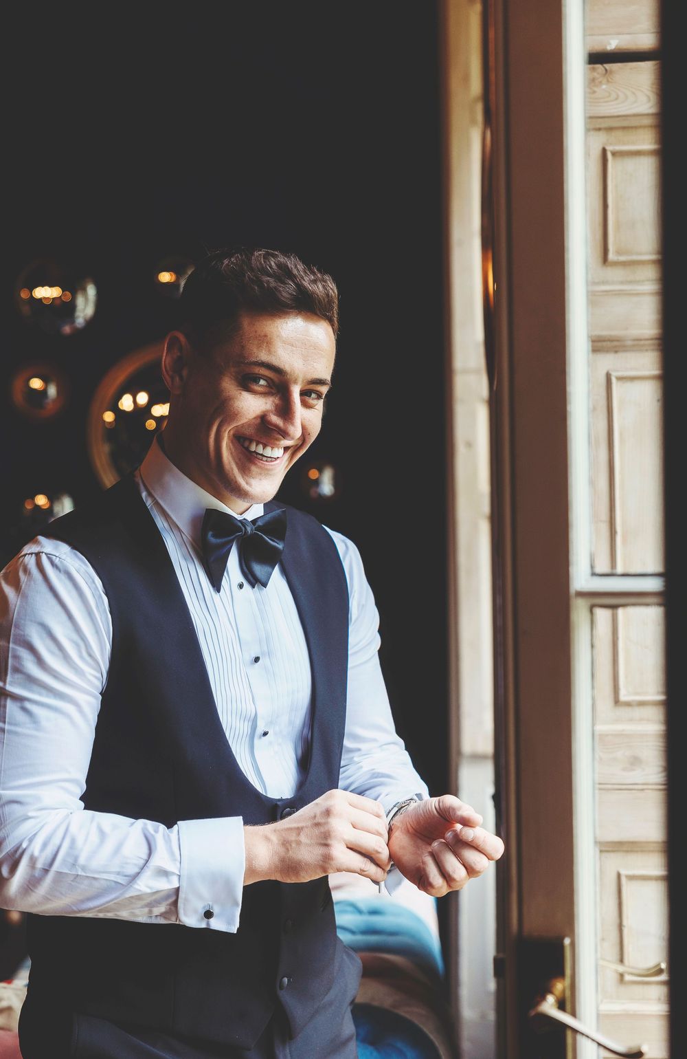 Groom in formal attire with black vest and bowtie smiling while looking at his watch indoors.