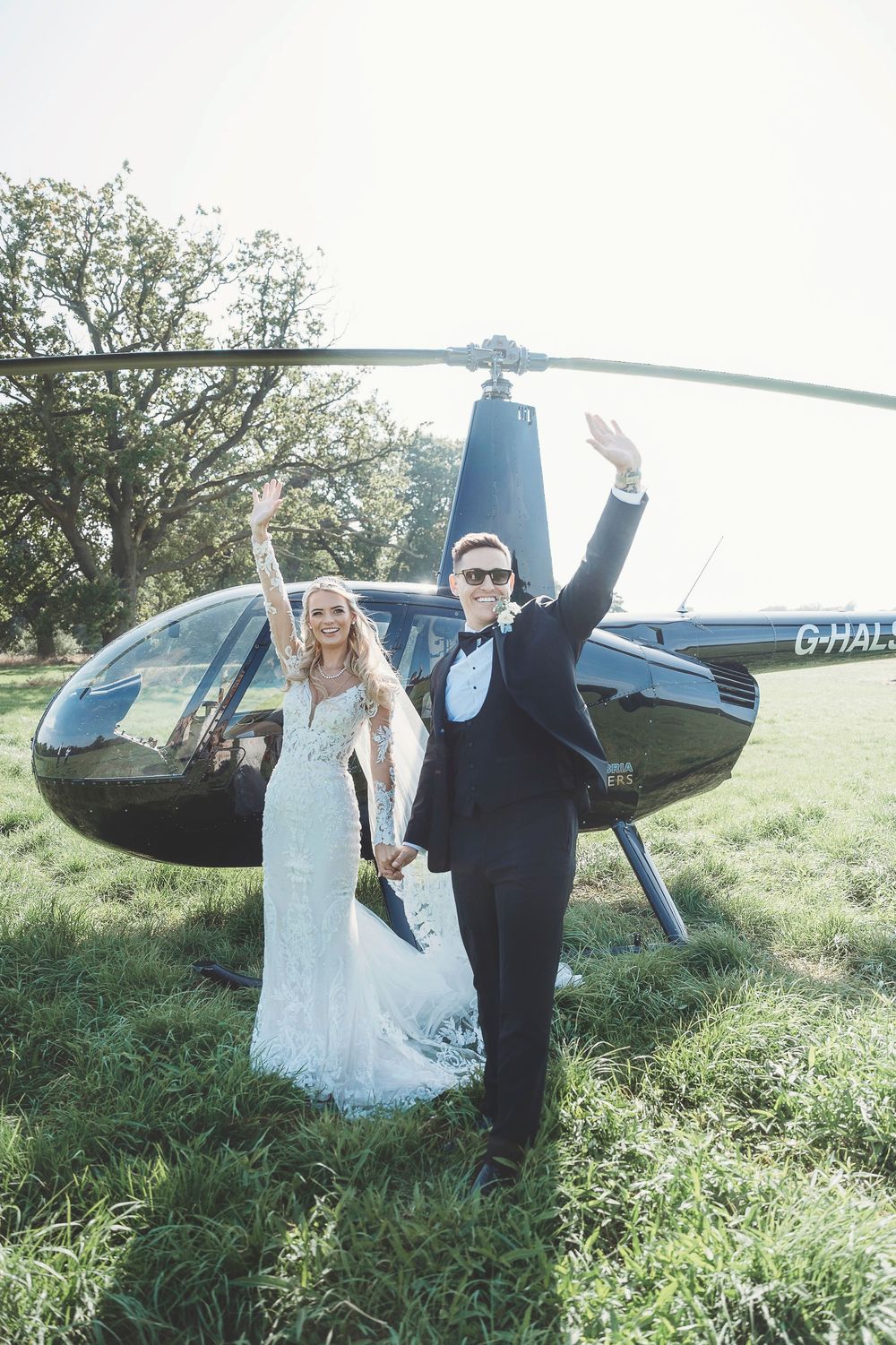 Celebratory wedding couple poses in a grassy field with a dark blue helicopter on a sunny day.