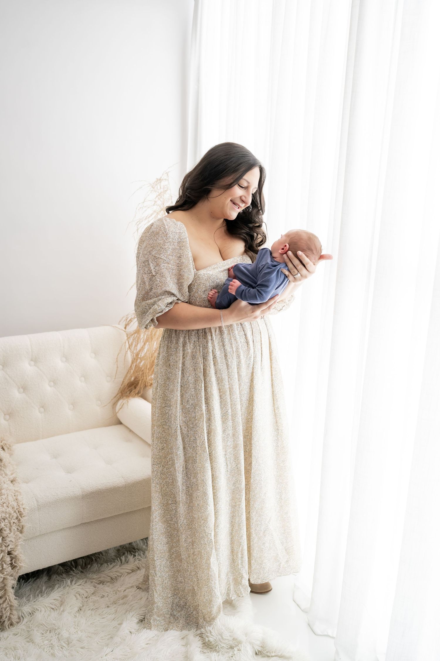 A person wearing a cream-colored lace dress cradles a newborn baby while sitting on a white tufted sofa in a bright room.