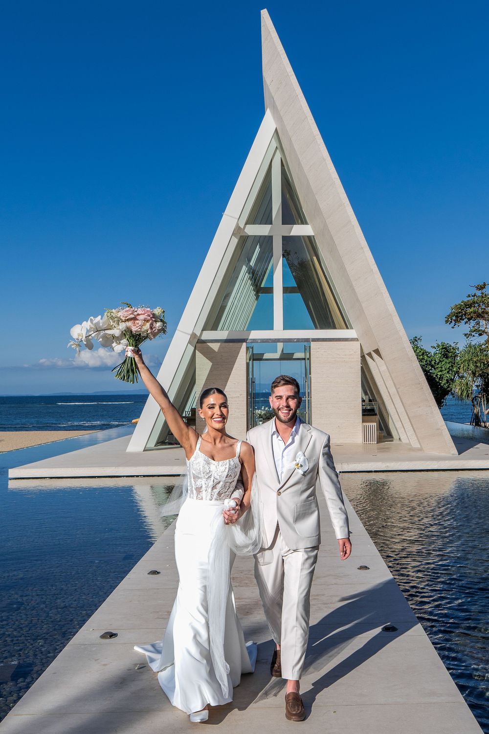 A newly married couple celebrates in front of a modern A-frame chapel with ocean views at Conrad Bali Resort.
