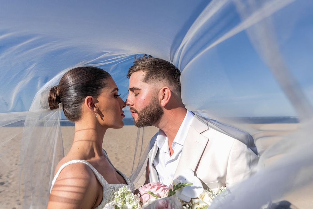 Newlywed couple kisses under flowing white veil with bright blue sky in background during outdoor wedding ceremony at Conrad Bali Resort.