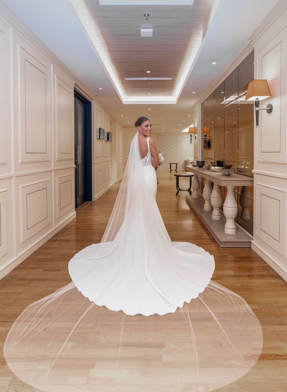 A wedding dress with flowing veil displayed along an elegant hallway with wood floors and white walls.