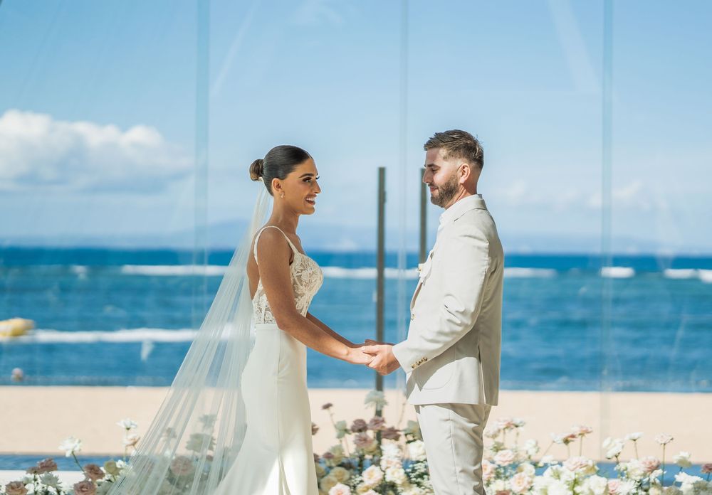 A romantic beach wedding ceremony with a couple holding hands against a backdrop of blue ocean and sky.